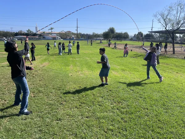 A teacher and students play jump rope on a grassy school field on a sunny day.