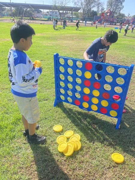 Two children play a giant Connect Four game on a grassy school playground on a sunny day.