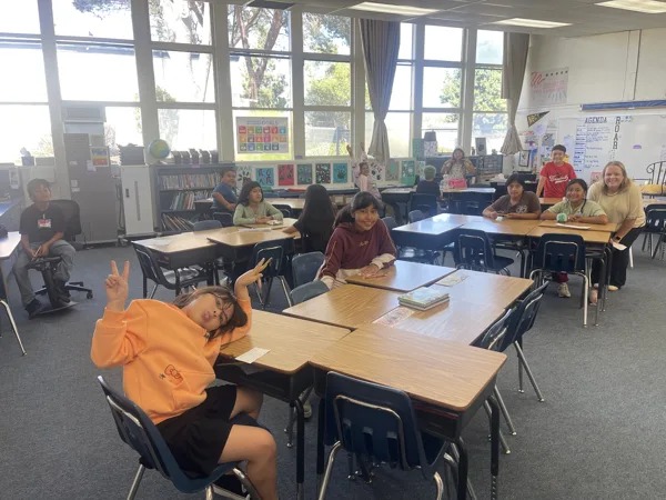 A group of elementary school students sit at desks in a bright classroom while a teacher smiles nearby, and a student in an orange hoodie poses with a peace sign toward the camera.