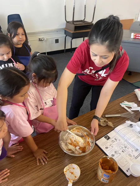 A woman in a red shirt helps young children mix ingredients in a bowl during a cooking activity in a classroom.