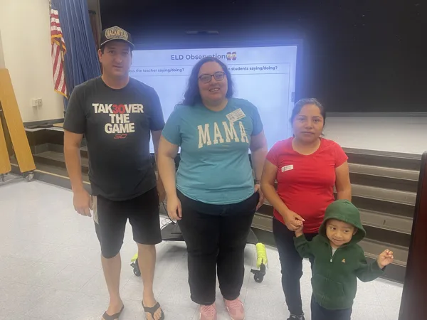 Three adults and a young child stand together in front of a presentation screen displaying an ELD Observations slide in what appears to be a school auditorium or meeting room.