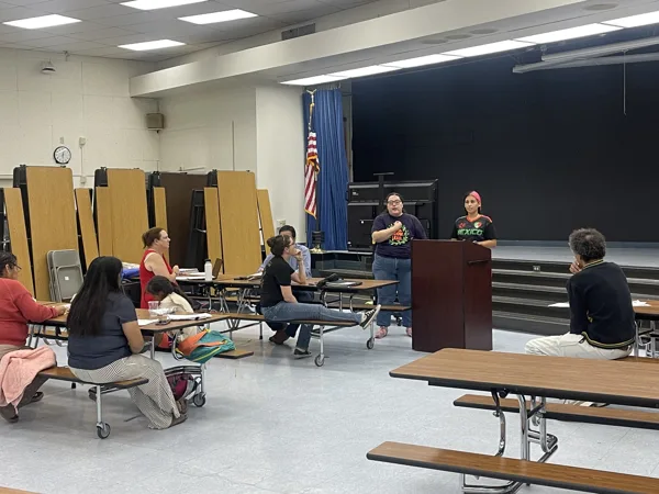 A woman speaks at a podium while a small group of adults sit at cafeteria tables listening in a school gymnasium or lunchroom.