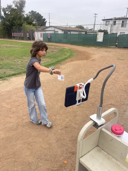 A child holds a QR code card up to a tablet mounted on a stand at an outdoor school playground.