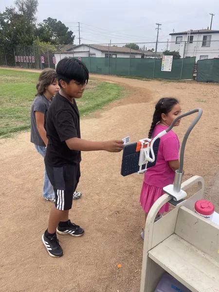 Three children stand on a school playground dirt field, with one boy holding a tablet device mounted in a holder near a cart.