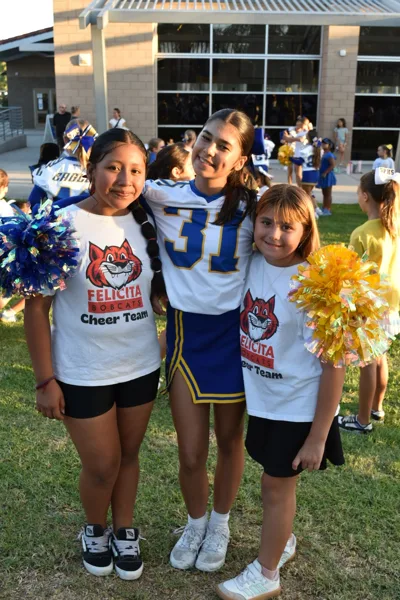 Three girls pose together outdoors at a school sports event, two wearing Felicita Bobcats Cheer Team shirts and one wearing a blue and white number 31 cheerleading uniform, with pom-poms in hand.