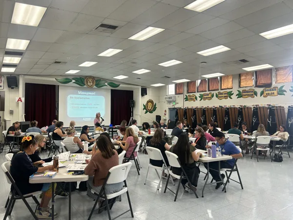 A group of adults sit at tables in a school cafeteria during a Kagan professional development training, with a presenter speaking at the front near a projected slide titled "Management.