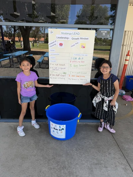 Two young girls stand outside a classroom holding a growth mindset poster titled "Mustangs LEAD" next to a blue bucket labeled "Rm 11.