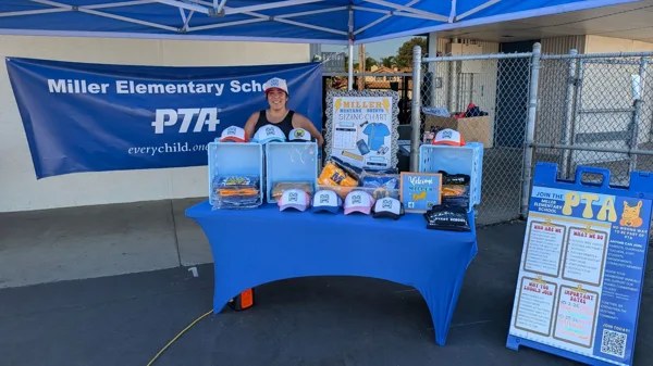 A smiling woman stands behind a blue-draped table selling hats and spirit wear at a Miller Elementary School PTA booth outdoors.