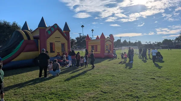 Children and adults gather around two inflatable bounce houses on a grassy field at an outdoor community event on a sunny day.