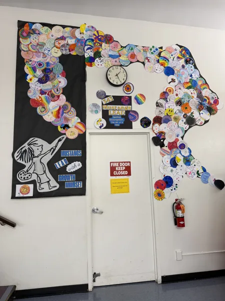 A school hallway wall displays hundreds of colorful student-made dot artworks arranged in a large arc around a fire door, alongside a banner reading "Mustangs LEAD with a Growth Mindset.