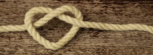 A cream-colored rope is tied in a heart-shaped knot on a weathered wooden surface.