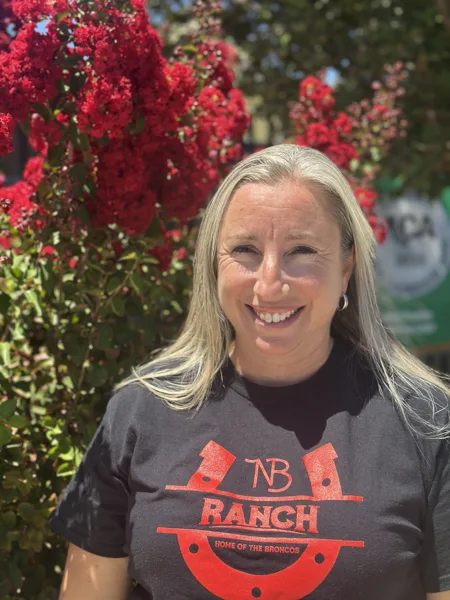 Principal Power smiling with long blonde hair wearing a dark NB Ranch Home of the Broncos t-shirt stands outdoors in front of a blooming red crape myrtle tree.