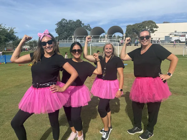 Four women wearing matching black tops and pink tutus flex their arms and pose on a school sports field during an outdoor event.