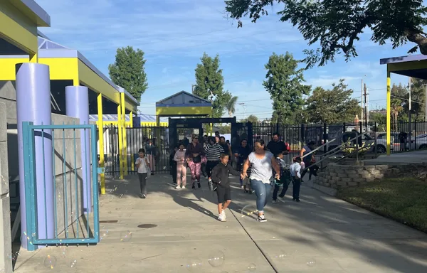 Children and adults walk out through the gated entrance of a school building on a sunny day.