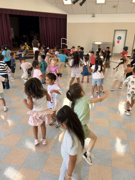 A large group of elementary school children dance together on a checkered tile floor in a school cafeteria or multipurpose room with a stage in the background.