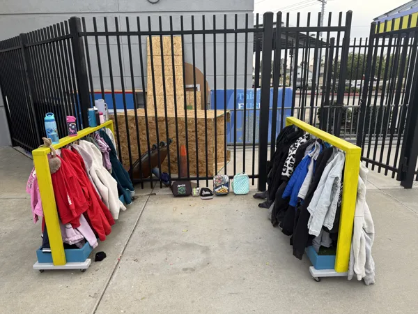 Two yellow clothing racks holding jackets and sweatshirts stand on a sidewalk outside a black metal fence, with lunch boxes and water bottles placed on the ground nearby.