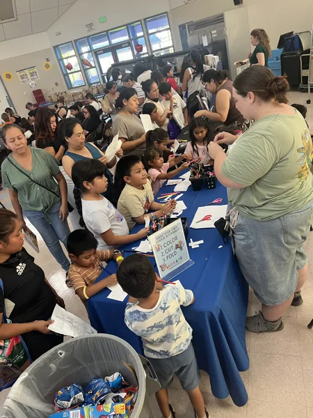 Children and adults gather around a blue-draped table at a busy indoor community event to make their own Alebrijes bookmarks by coloring, cutting, and folding paper.