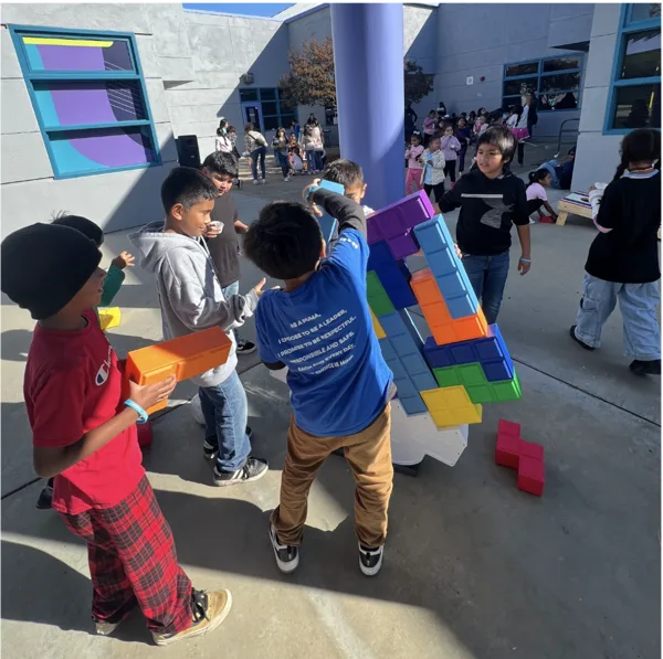 Children play with large colorful Tetris-shaped building blocks in a school courtyard during recess.