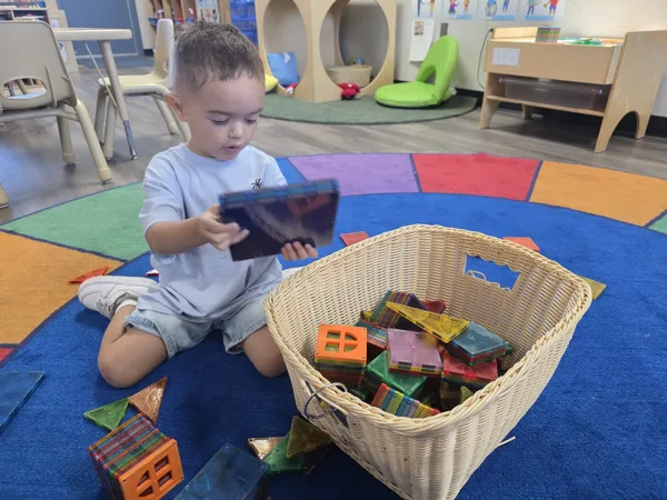 A young boy sits on a colorful classroom rug, holding a tablet, with a basket of magnetic tiles beside him.