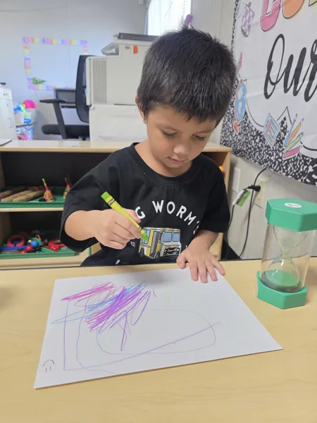 A young boy draws on white paper with a yellow crayon at a classroom table, with a green sand timer beside him.