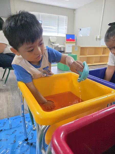 A young boy plays at a water table in a classroom, pouring water from a small toy into a yellow bin.