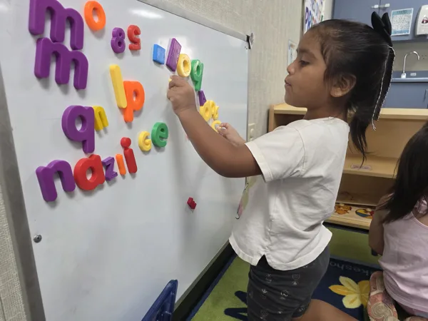 A young girl places colorful magnetic letters on a whiteboard in a classroom.