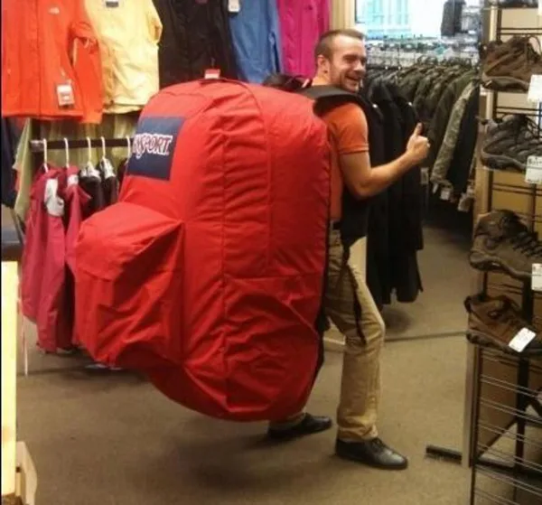 A man tries on an oversized red JanSport backpack in a sporting goods store, giving a thumbs up.