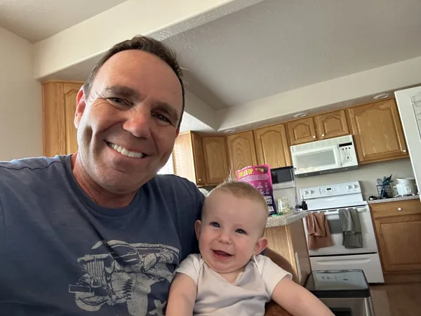 A smiling middle-aged man holds a happy baby in a home kitchen.