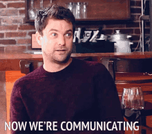 A man in a dark red sweater sits at a bar with a wine glass nearby, with the caption "NOW WE'RE COMMUNICATING" at the bottom of the image.
