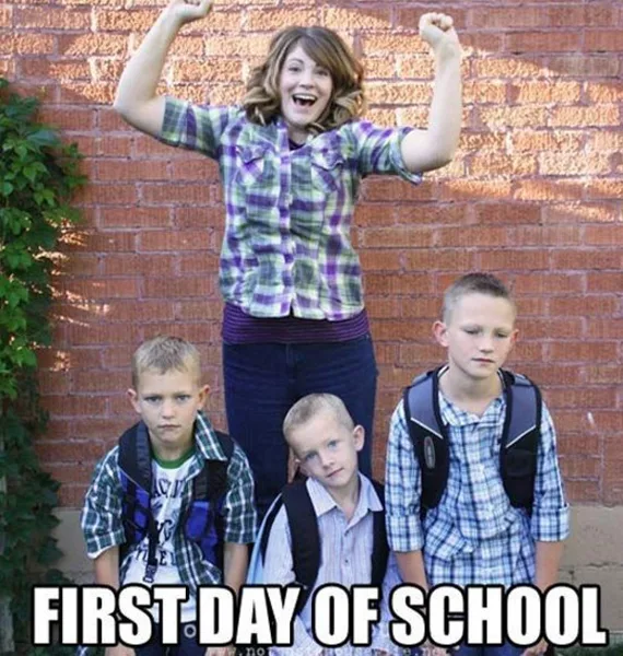 A woman cheers with raised fists while three young boys wearing backpacks look unenthusiastic in front of a brick wall on the first day of school.