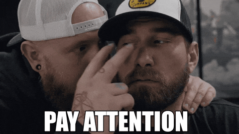 Two bearded men, one wearing a snapback cap, share an emotional moment as one gestures near the other's face, with the text "PAY ATTENTION" displayed at the bottom.