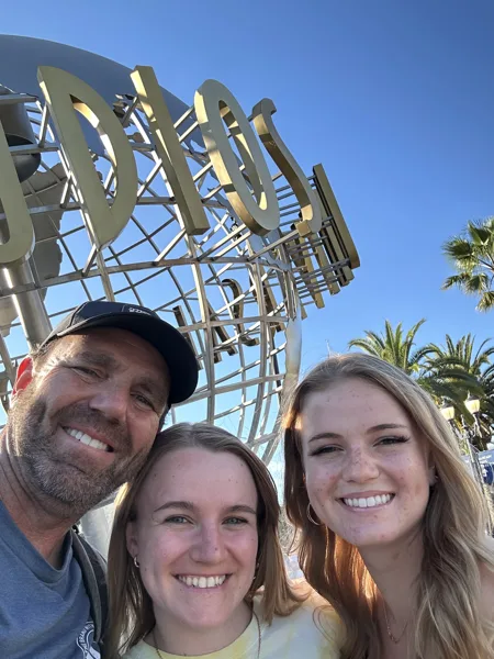 Three smiling people take a selfie in front of the Universal Studios globe sculpture on a sunny day.