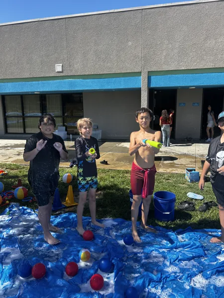 Three children stand on a wet blue tarp with foam balls during an outdoor water play activity beside a gray building on a sunny day.