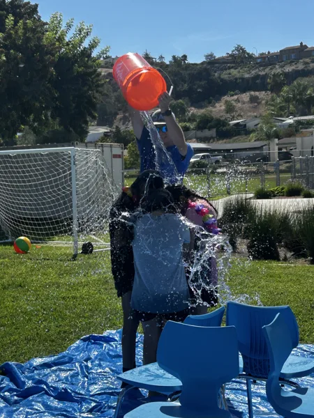 A person pours a large orange bucket of water over another person standing on a blue tarp in a sunny outdoor park with a soccer goal in the background.