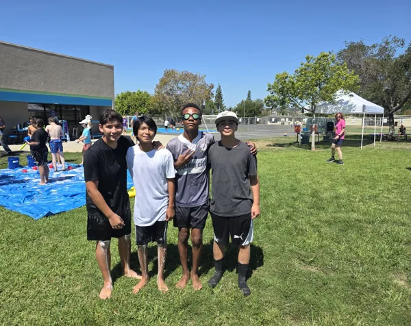 Four young teenagers pose together on a grass field at an outdoor summer water day event, with a slip-and-slide tarp visible in the background.