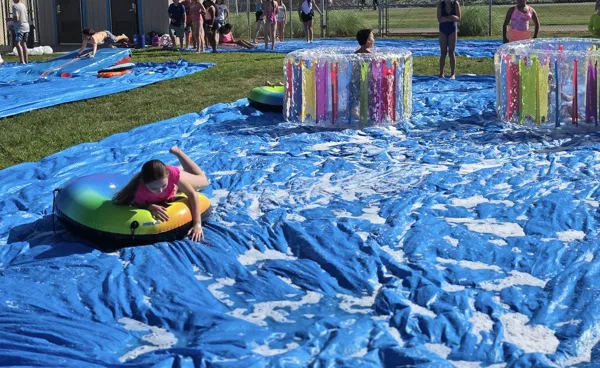 Children play on a soapy blue tarp slip-and-slide with inflatable tubes and small pools in a grassy outdoor area on a sunny day.