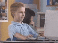 A young boy sits at a desktop computer in a classroom, smiling while using the mouse.