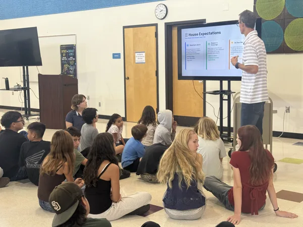 A teacher stands beside a screen displaying House Expectations while a group of middle school students sit on the floor of a school cafeteria listening to the presentation.