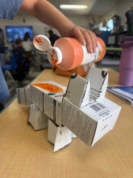 A child pours orange paint onto a small animal sculpture made from recycled cardboard boxes on a classroom desk.