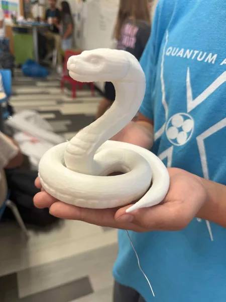 A student in a blue Quantum shirt holds a white 3D-printed snake sculpture in what appears to be a classroom or makerspace.
