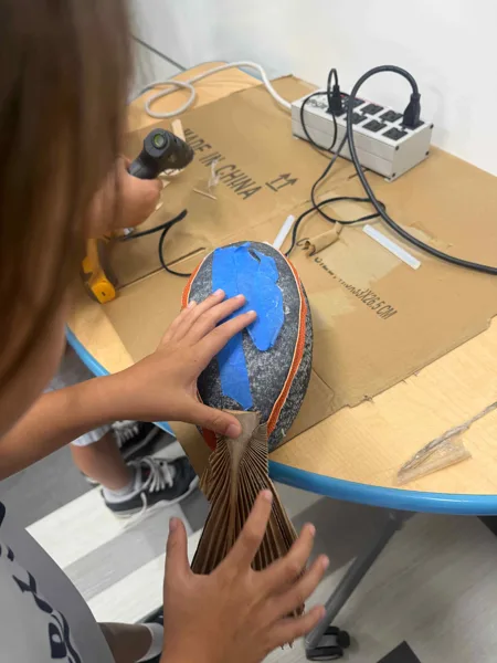 A child holds a handmade fish sculpture decorated with blue tape and cardboard fins over a worktable covered with craft supplies and a power strip.
