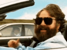 A bearded man wearing sunglasses sits in the driver's seat of a convertible car on a sunny day.