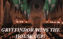 A large group of students in robes celebrates inside a grand candle-lit dining hall decorated with green banners, with text reading GRYFFINDOR WINS THE HOUSE CUP!