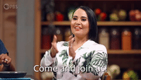 A smiling woman in a floral blouse claps her hands in what appears to be a television cooking show studio, with the caption "Come and join us" displayed on screen.