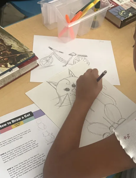 A child draws a cat on paper at a desk, using a pencil and following a "How to Draw a Cat" instruction guide, with other drawings and art supplies nearby.
