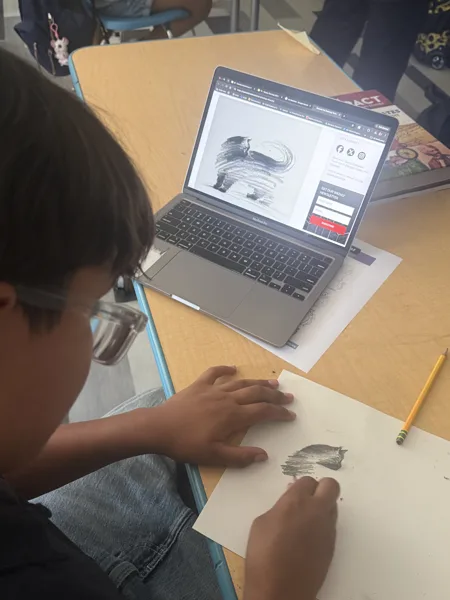 A young child wearing glasses draws on paper at a school desk while referencing a black-and-white artwork displayed on a MacBook Pro laptop.