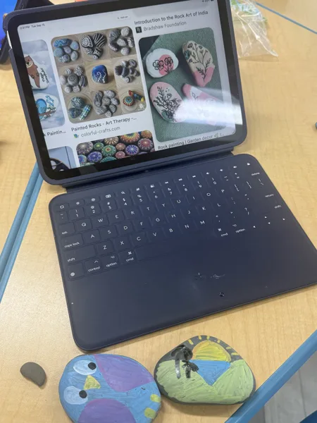 An iPad with a keyboard case displays rock painting search results next to two hand-painted rocks on a wooden desk.