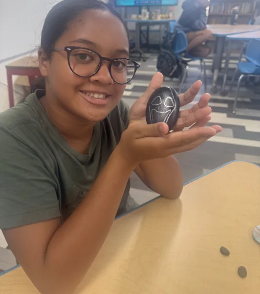 A young woman wearing glasses holds up a black painted rock decorated with a white ghost face while sitting at a table in what appears to be a school or library setting.