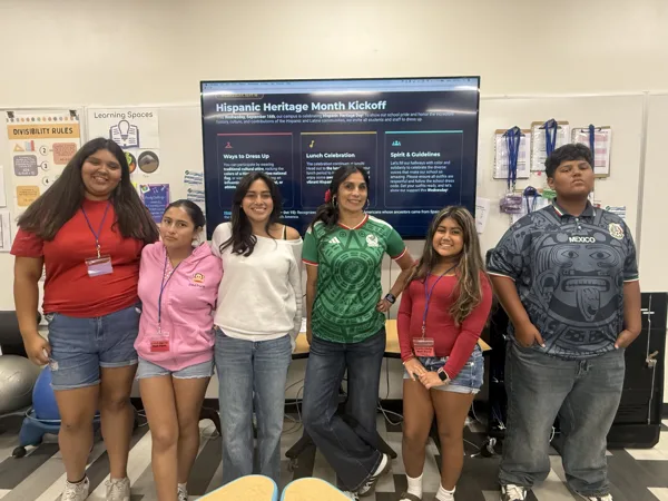 Six people pose together in a classroom in front of a screen displaying a Hispanic Heritage Month Kickoff announcement.