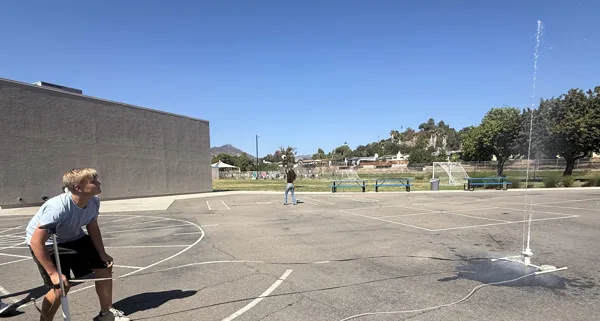 A boy pumps air into a water rocket on a school blacktop as it launches a stream of water into the air, with a soccer field and hills visible in the background.